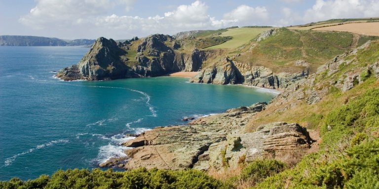 Beach in Devon, UK. Sandy shore with clear blue water, surrounded by cliffs and greenery.